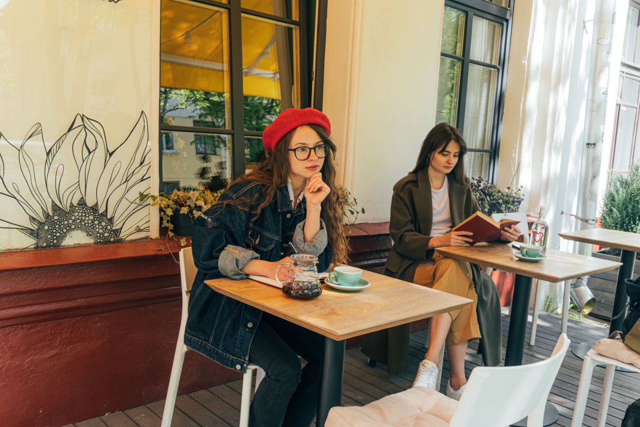 Zwei Menschen sitzen mit Kaffee und Notizbuch an Tischen eines hellen Cafés